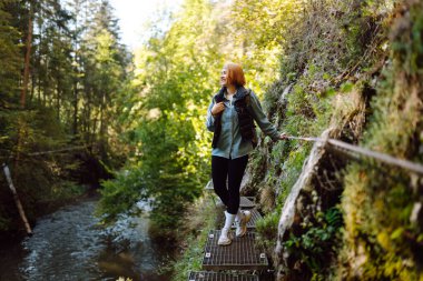 Sırt çantalı güzel bir bayan yürüyüşçü derenin yanındaki metal yürüyüş yolunda duruyor. Genç bir kadın güneşli ormanda vahşi doğanın tadını çıkarıyor. Doğa konsepti, yürüyüş. Etkin yaşam biçimi.