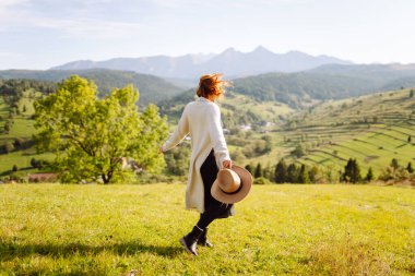Güneşli bir günde yeşil tepelerde eğlenen güzel bir kadının arka görüntüsü. Şapkalı bir gezgin gün batımında dağ manzarasının tadını çıkarıyor. Eğlence, seyahat, rahatlama kavramı.