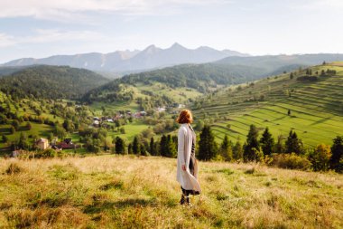 Güneşli bir günde yeşil tepelerde eğlenen güzel bir kadının arka görüntüsü. Şapkalı bir gezgin gün batımında dağ manzarasının tadını çıkarıyor. Eğlence, seyahat, rahatlama kavramı.
