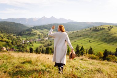 Güneşli bir günde yeşil tepelerde eğlenen güzel bir kadının arka görüntüsü. Şapkalı bir gezgin gün batımında dağ manzarasının tadını çıkarıyor. Eğlence, seyahat, rahatlama kavramı.