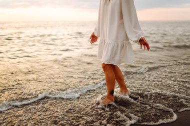 Gün batımında sahilde yürüyen bir kadının bacaklarının yakın çekimi. Genç bir kadın deniz manzarasının tadını çıkarıyor, sahilde yürüyor. Doğa, rahatlama konsepti.