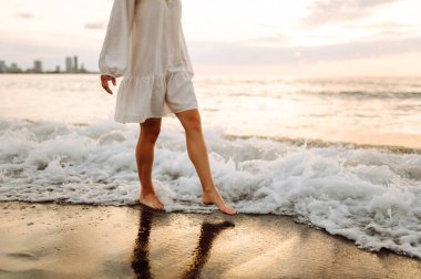 Gün batımında sahilde yürüyen bir kadının bacaklarının yakın çekimi. Genç bir kadın deniz manzarasının tadını çıkarıyor, sahilde yürüyor. Doğa, rahatlama konsepti.