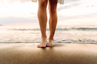 Gün batımında sahilde yürüyen bir kadının bacaklarının yakın çekimi. Genç bir kadın deniz manzarasının tadını çıkarıyor, sahilde yürüyor. Doğa, rahatlama konsepti.