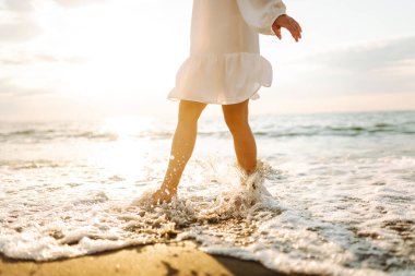 Gün batımında sahilde yürüyen bir kadının bacaklarının yakın çekimi. Genç bir kadın deniz manzarasının tadını çıkarıyor, sahilde yürüyor. Doğa, rahatlama konsepti.