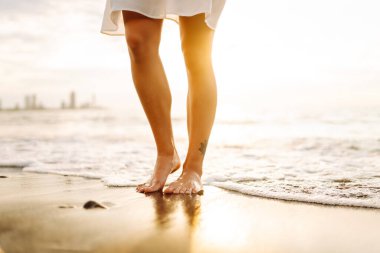 Gün batımında sahilde yürüyen bir kadının bacaklarının yakın çekimi. Genç bir kadın deniz manzarasının tadını çıkarıyor, sahilde yürüyor. Doğa, rahatlama konsepti.