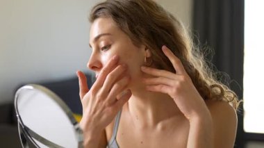 Genç bir kadın sıcak bir odada küçük bir aynaya bakarken yüzüne dokunuyor. Güzel bir kadın doğal güzellikten zevk alır ve cilt bakımı yapar. Dermatoloji, doğallık ve güzellik kavramları.