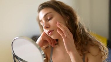 Genç bir kadın sıcak bir odada küçük bir aynaya bakarken yüzüne dokunuyor. Güzel bir kadın doğal güzellikten zevk alır ve cilt bakımı yapar. Dermatoloji, doğallık ve güzellik kavramları.
