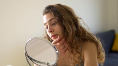 Genç bir kadın sıcak bir odada küçük bir aynaya bakarken yüzüne dokunuyor. Güzel bir kadın doğal güzellikten zevk alır ve cilt bakımı yapar. Dermatoloji, doğallık ve güzellik kavramları.