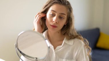 Genç bir kadın sıcak bir odada küçük bir aynaya bakarken yüzüne dokunuyor. Güzel bir kadın doğal güzellikten zevk alır ve cilt bakımı yapar. Dermatoloji, doğallık ve güzellik kavramları.