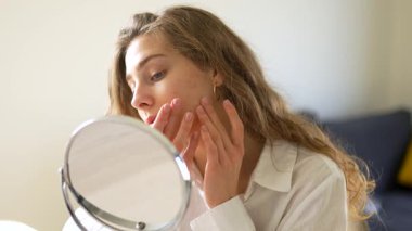 Genç bir kadın sıcak bir odada küçük bir aynaya bakarken yüzüne dokunuyor. Güzel bir kadın doğal güzellikten zevk alır ve cilt bakımı yapar. Dermatoloji, doğallık ve güzellik kavramları.