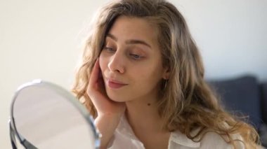 Genç bir kadın sıcak bir odada küçük bir aynaya bakarken yüzüne dokunuyor. Güzel bir kadın doğal güzellikten zevk alır ve cilt bakımı yapar. Dermatoloji, doğallık ve güzellik kavramları.