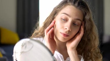 Genç bir kadın sıcak bir odada küçük bir aynaya bakarken yüzüne dokunuyor. Güzel bir kadın doğal güzellikten zevk alır ve cilt bakımı yapar. Dermatoloji, doğallık ve güzellik kavramları.