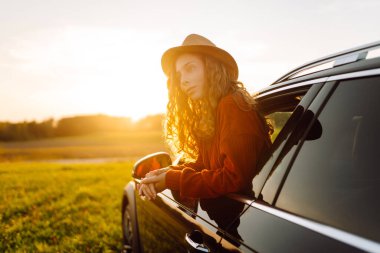 Şapkalı hoş bir kadın arabanın camından dışarı bakıyor, güneşli havanın tadını çıkarıyor. Güzel kadın pencereden dışarı bakarken kendini özgür hissediyor. Konsept: araba macerası, özgürlük, hafta sonu. Etkin yaşam biçimi.