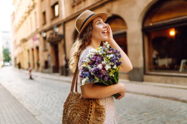 Şapkalı neşeli kadın, güneşli cadde boyunca kır çiçekleriyle geziyor. Güzel bir kadın buketi tutuyor ve şehir atmosferinin tadını çıkarıyor. Yürüme, dinlenme ve turizm için kavramsal kavramlar.
