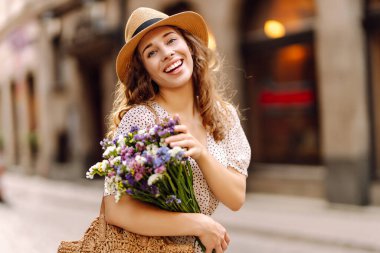 Şapkalı neşeli kadın, güneşli cadde boyunca kır çiçekleriyle geziyor. Güzel bir kadın buketi tutuyor ve şehir atmosferinin tadını çıkarıyor. Yürüme, dinlenme ve turizm için kavramsal kavramlar.