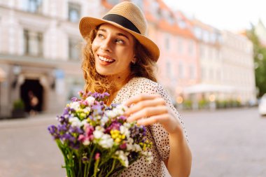 Şapkalı neşeli kadın, güneşli cadde boyunca kır çiçekleriyle geziyor. Güzel bir kadın buketi tutuyor ve şehir atmosferinin tadını çıkarıyor. Yürüme, dinlenme ve turizm için kavramsal kavramlar.