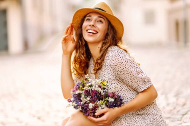 Şapkalı neşeli kadın, güneşli cadde boyunca kır çiçekleriyle geziyor. Güzel bir kadın buketi tutuyor ve şehir atmosferinin tadını çıkarıyor. Yürüme, dinlenme ve turizm için kavramsal kavramlar.