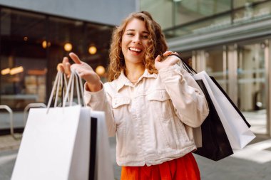 Parlak elbiseli neşeli bir kadın alışveriş torbalarıyla şehir merkezinde duruyor. Genç bir kadın alışveriş sonrası çantalarla güneşli bir günün tadını çıkarır. Konsept: indirim, alışveriş. Yaşam biçimi.