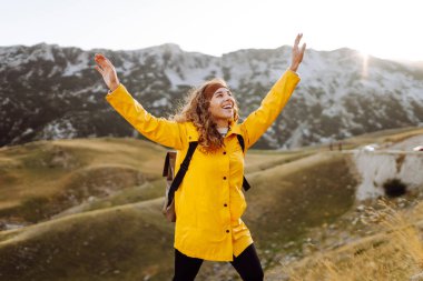 Sırt çantalı, parlak sarı yağmurluklu neşeli bir kadın dağ manzarasının tadını çıkarıyor. Bir yürüyüşçü dağ yolunda yürürken kendini özgür hisseder. Etkin yaşam biçimi.