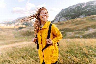 Sırt çantalı, parlak sarı yağmurluklu neşeli bir kadın dağ manzarasının tadını çıkarıyor. Bir yürüyüşçü dağ yolunda yürürken kendini özgür hisseder. Etkin yaşam biçimi.