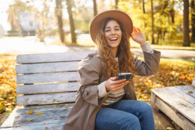 Şapkalı neşeli bir kadının portresi, telefon kullanması, sonbahar parkında bankta oturması. Güzel bir kadın, ahşap bankta telefon kullanarak güneşli bir günün tadını çıkarır. Blog yazma, gevşeme ve mevsimlik kavramı.