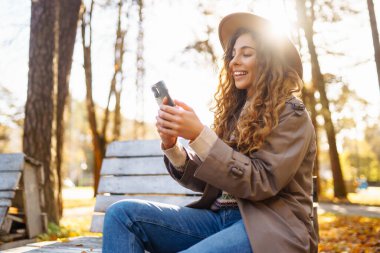 Şapkalı neşeli bir kadının portresi, telefon kullanması, sonbahar parkında bankta oturması. Güzel bir kadın, ahşap bankta telefon kullanarak güneşli bir günün tadını çıkarır. Blog yazma, gevşeme ve mevsimlik kavramı.