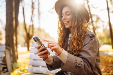Şapkalı neşeli bir kadının portresi, telefon kullanması, sonbahar parkında bankta oturması. Güzel bir kadın, ahşap bankta telefon kullanarak güneşli bir günün tadını çıkarır. Blog yazma, gevşeme ve mevsimlik kavramı.