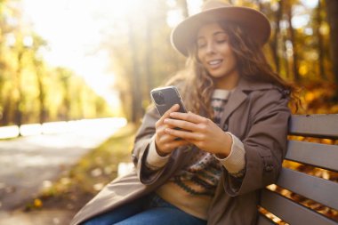 Şapkalı neşeli bir kadının portresi, telefon kullanması, sonbahar parkında bankta oturması. Güzel bir kadın, ahşap bankta telefon kullanarak güneşli bir günün tadını çıkarır. Blog yazma, gevşeme ve mevsimlik kavramı.