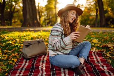 Genç bir kadın elinde bir kitapla sonbahar otlağında kırmızı bir battaniyeye oturur. Şapkalı gülümseyen bir kadın kitap okur ve parkın doğal güzelliğinden zevk alır. Kavramları: rahatlama, doğa, okuma.