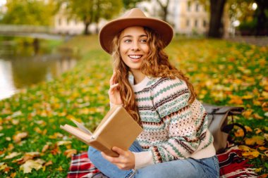 Genç bir kadın elinde bir kitapla sonbahar otlağında kırmızı bir battaniyeye oturur. Şapkalı gülümseyen bir kadın kitap okur ve parkın doğal güzelliğinden zevk alır. Kavramları: rahatlama, doğa, okuma.