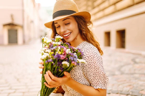 Şapkalı neşeli kadın, güneşli cadde boyunca kır çiçekleriyle geziyor. Güzel bir kadın buketi tutuyor ve şehir atmosferinin tadını çıkarıyor. Yürüme, dinlenme ve turizm için kavramsal kavramlar.