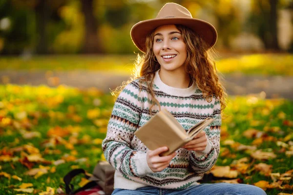 Genç bir kadın elinde bir kitapla sonbahar otlağında kırmızı bir battaniyeye oturur. Şapkalı gülümseyen bir kadın kitap okur ve parkın doğal güzelliğinden zevk alır. Kavramları: rahatlama, doğa, okuma.