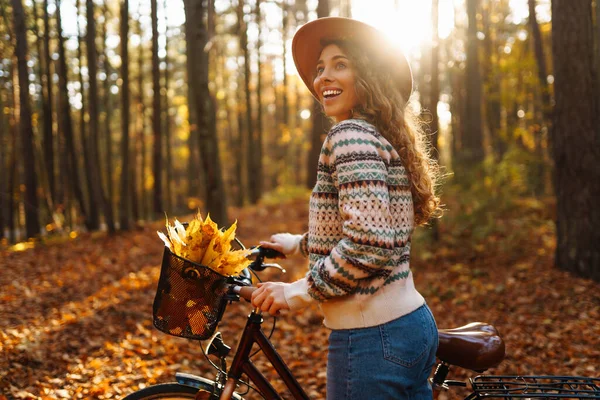 Sonbahar ormanında şapkalı, bisikletli gülümseyen bir kadın. Genç bir kadın bisiklet sürüyor ve gün batımında doğanın tadını çıkarıyor. Rahatlama kavramı, hafta sonları, eğlence.