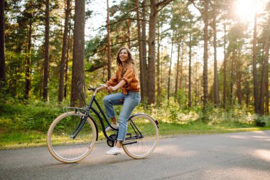 Neşeli bir kadın güneşli bir ormanda bisiklet sürüyor. Güzel bir kadın doğal çevreden zevk alır ve gün batımında bisiklete biner. Hafta sonu ve eğlence konsepti.