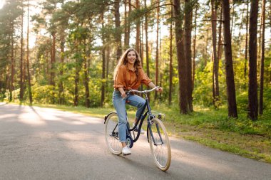 Neşeli bir kadın güneşli bir ormanda bisiklet sürüyor. Güzel bir kadın doğal çevreden zevk alır ve gün batımında bisiklete biner. Hafta sonu ve eğlence konsepti.