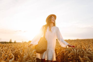 Buğday tarlasında genç bir kadın günbatımının tadını çıkarıyor. Beyaz elbiseli ve şapkalı neşeli bir kadın güneşli bir günde altın bir tarlada geziniyor. Güzellik ve doğa kavramı.