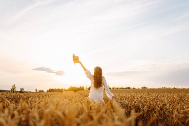 Buğday tarlasında genç bir kadın günbatımının tadını çıkarıyor. Beyaz elbiseli ve şapkalı neşeli bir kadın güneşli bir günde altın bir tarlada geziniyor. Güzellik ve doğa kavramı.