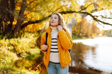 Sarı ceketli ve sırtında bir sırt çantası taşıyan güzel bir yürüyüşçü nehir boyunca bir yürüyüş yolu boyunca yürüyor. Genç kadın sonbahar manzarasının tadını çıkarıyor. Macera kavramları, mevsimlik.