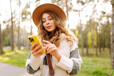 Güneşli bir parkta telefonla dolaşan şapkalı güzel bir kadının portresi. Telefonu olan genç bir turist açık havada güneşli bir günün tadını çıkarıyor. Blog yazma ve boş zaman kavramları.