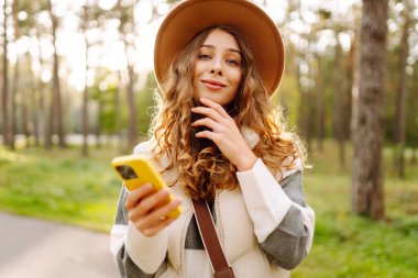 Güneşli bir parkta telefonla dolaşan şapkalı güzel bir kadının portresi. Telefonu olan genç bir turist açık havada güneşli bir günün tadını çıkarıyor. Blog yazma ve boş zaman kavramları.