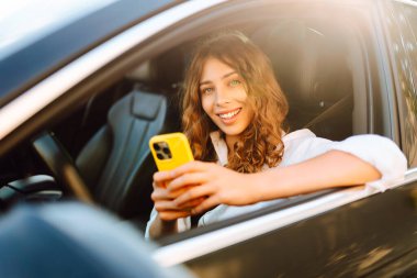 Telefonu olan gülümseyen bir kadın sürücü koltuğunda oturur ve pencereden dışarı bakar. Bir kadın sürücü, direksiyon başında güneşli bir günün tadını çıkarır. Ulaşım ve teknoloji kavramı.