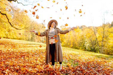 Mutlu bir kadın gün batımında bir sonbahar parkında eğleniyor. Gündelik giysiler içinde güzel bir kadın güneşli bir günde havaya yapraklar fırlatır. Yürüyüş kavramı, rahatlama..
