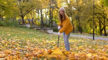 Genç bir kadın, uzun ağaçlarla çevrili bir parkta canlı turuncu ve sarı yapraklar tırmıklıyor. Sonbaharın parlak renkleri toprağı düzenlerken resim gibi bir sahne yaratıyor..