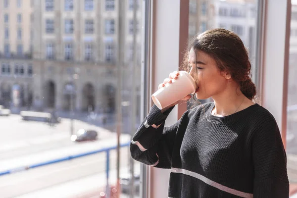 Dziewczyna pije kawę i stoi przez panoramiczne okno — Zdjęcie stockowe