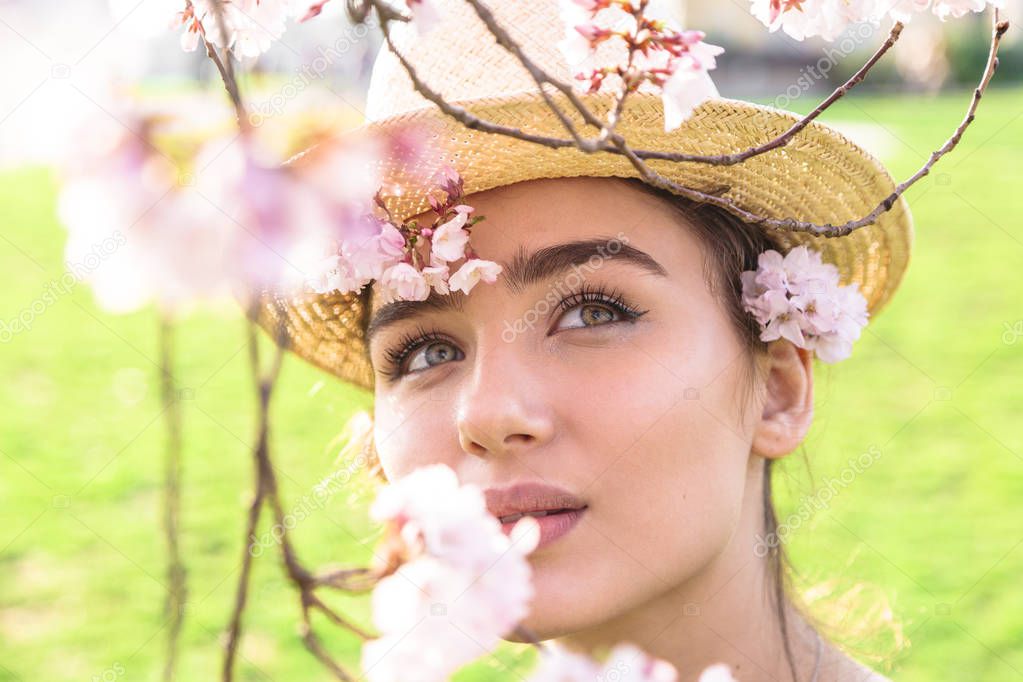Girl in straw hat dreamily looks at blossom branch