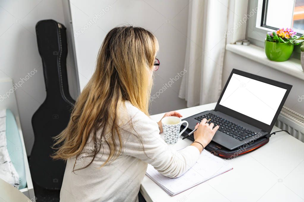 Girl works at laptop at table by window at home