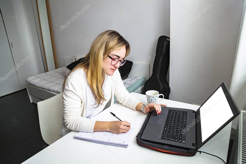 Girl works at laptop at table by window at home