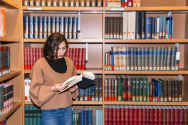 Kütüphanede kitaplıkların yanında duran genç kız.