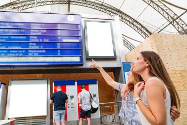 Kadın, tren istasyonundaki bir panoda arkadaşının programını gösteriyor.