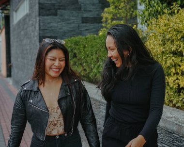 İki neşeli genç kadın, güneşli bir günde, yemyeşil bir çitin yanında, asfalt bir sokakta birlikte yürüyüp, kahkahalarla eğleniyorlar.